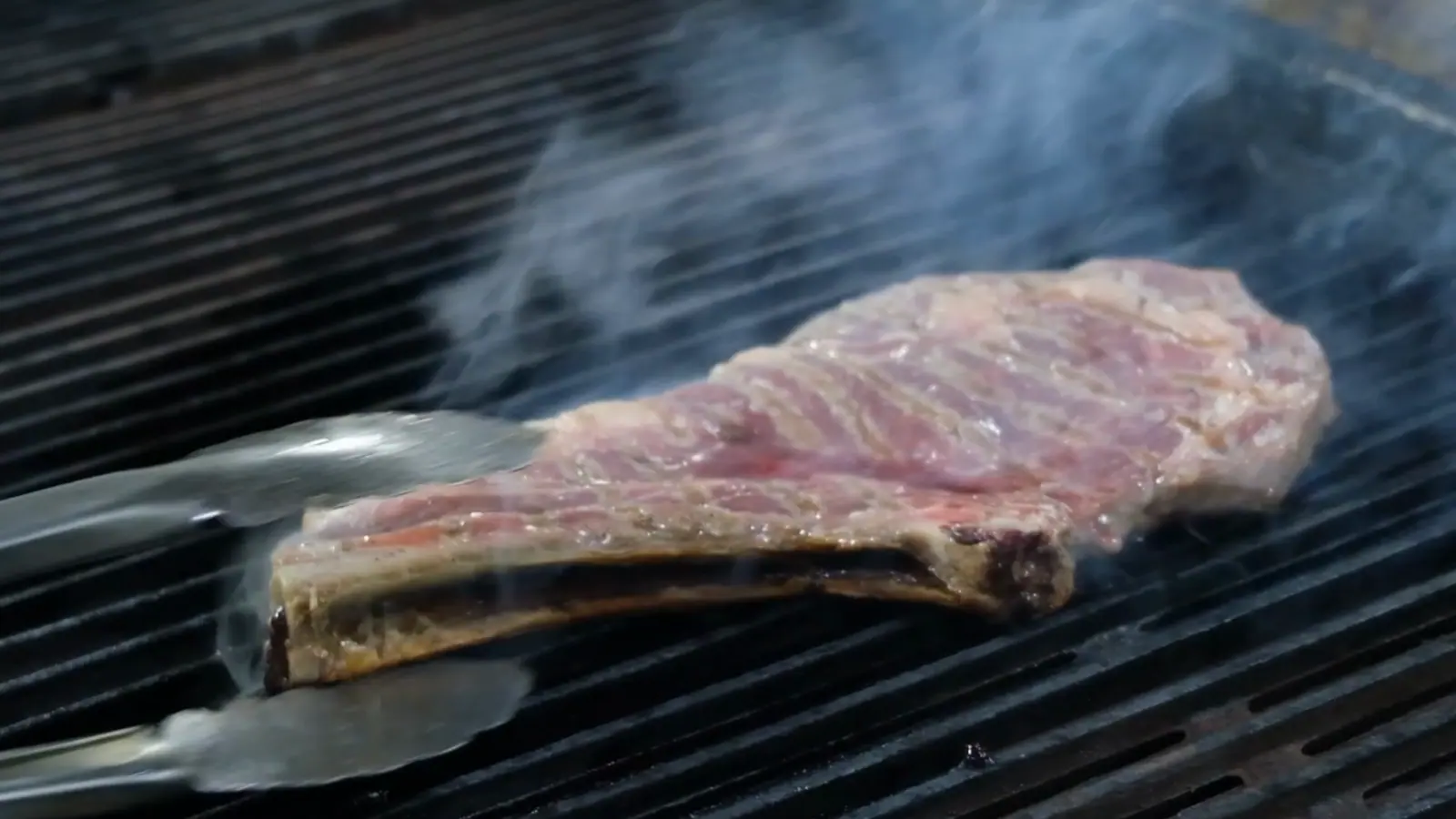 En grilltang vender kjøtt på grillristen mens røyk stiger opp.
