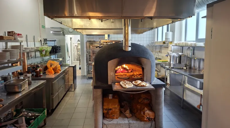 Åpen vedovn i restaurantkjøkkenet med flammer og flatbrød i og ved ovnsåpningen.